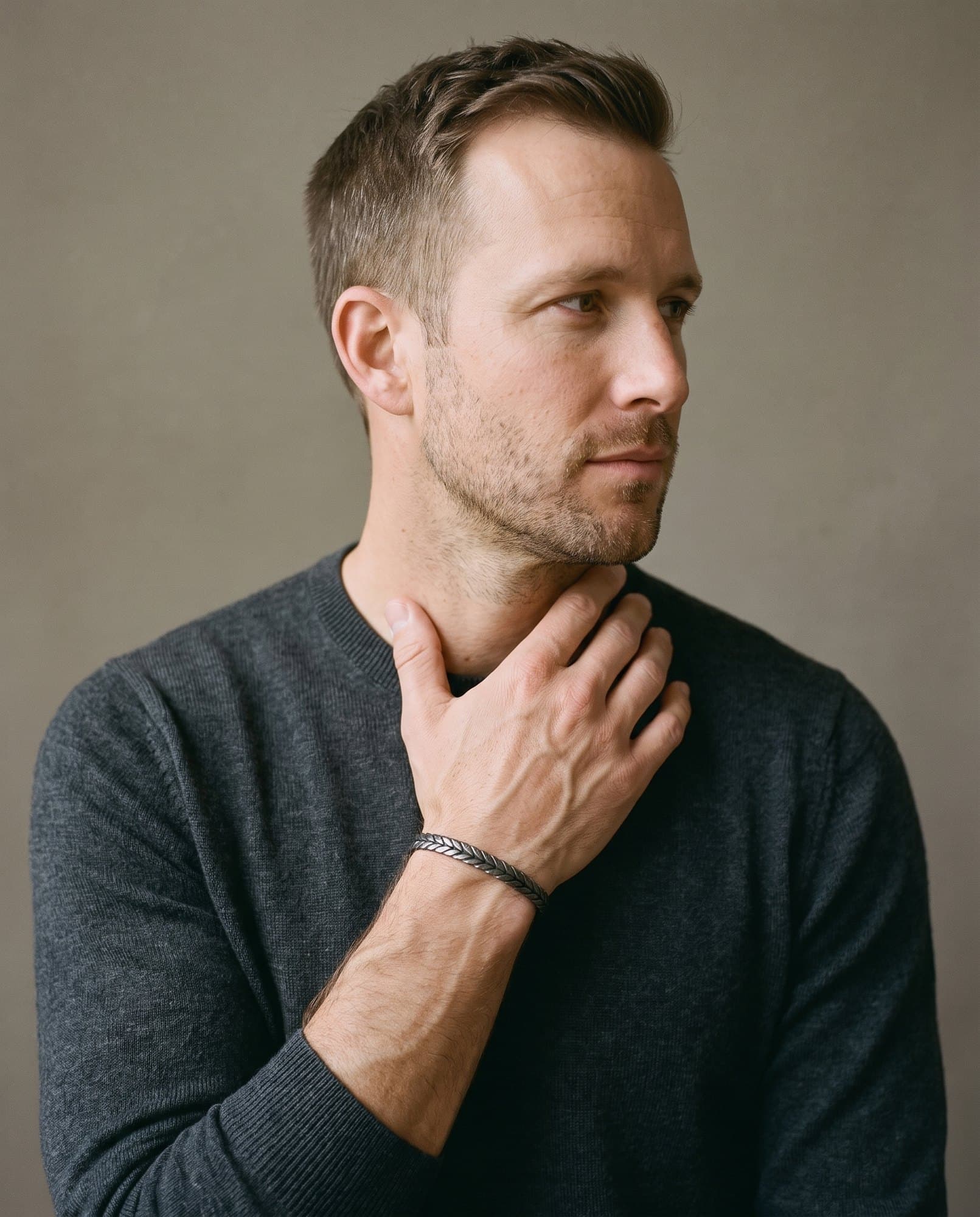 A man in a charcoal knit with his hand at his collarbone, the Amun Braid Cuff on his wrist.