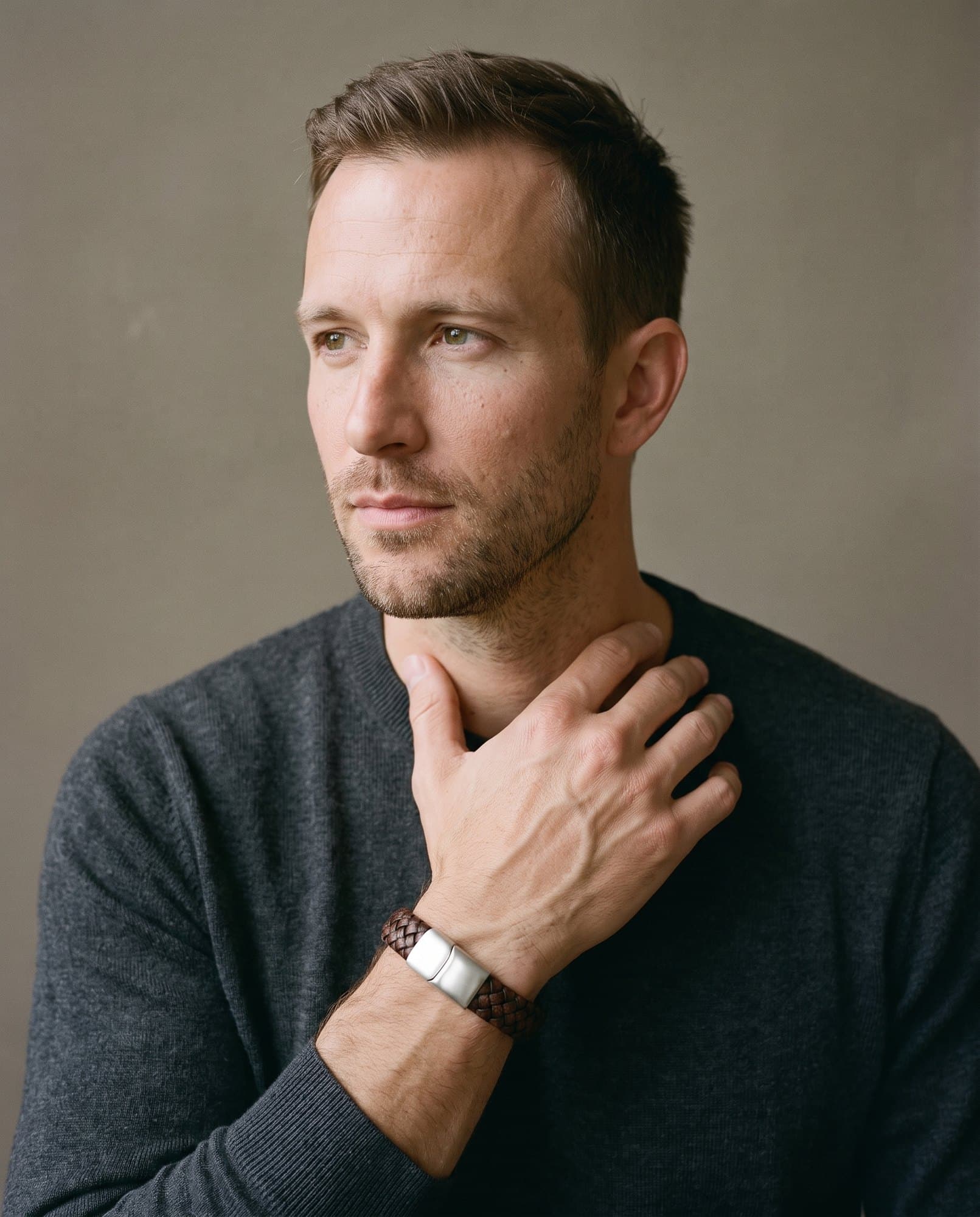 A man in a charcoal knit with his hand at his collarbone, the Broad Plait Cord on his wrist.