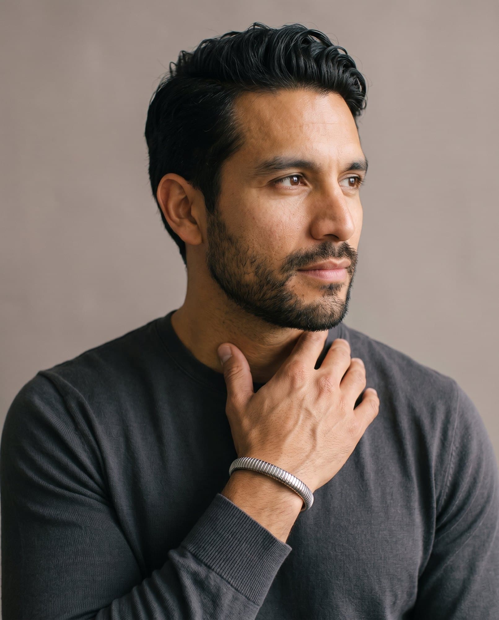 A man in a charcoal knit with his hand at his collarbone, the Coil Bangle on his wrist.