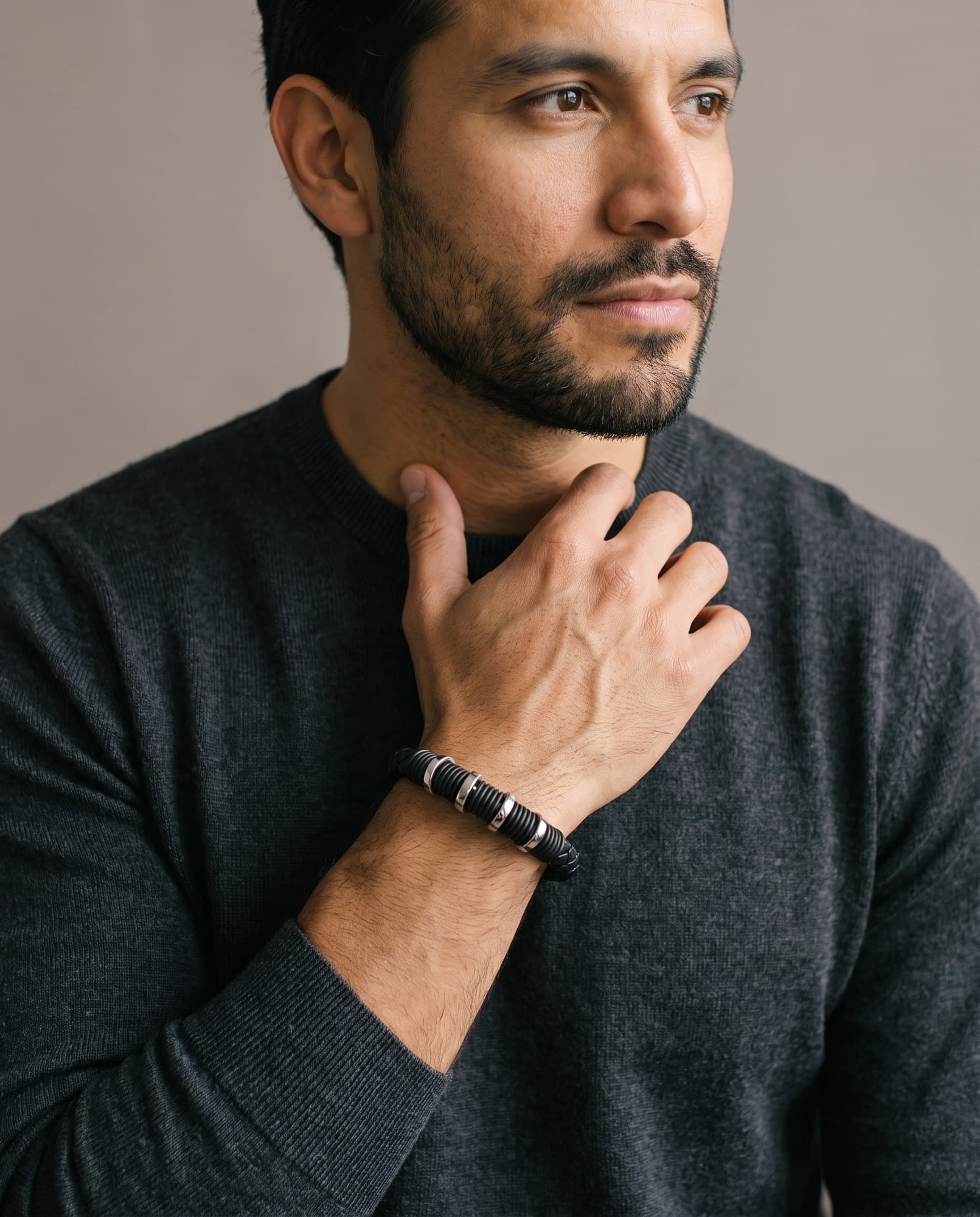 A man in a charcoal knit with his hand at his collarbone, the Coil Ring Cord on his wrist.
