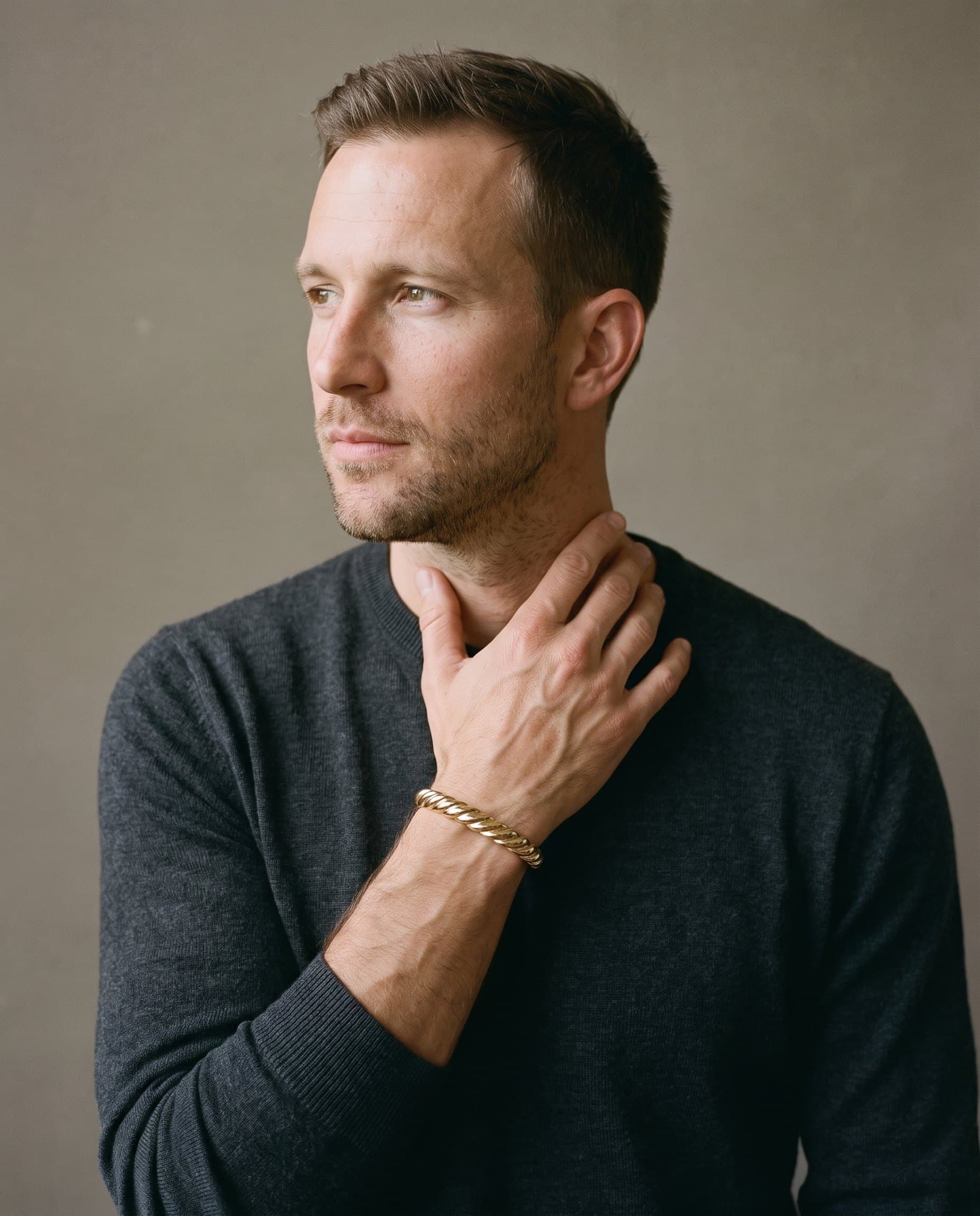 A man in a charcoal knit with his hand at his collarbone, the Giza Wheat Cuff on his wrist.