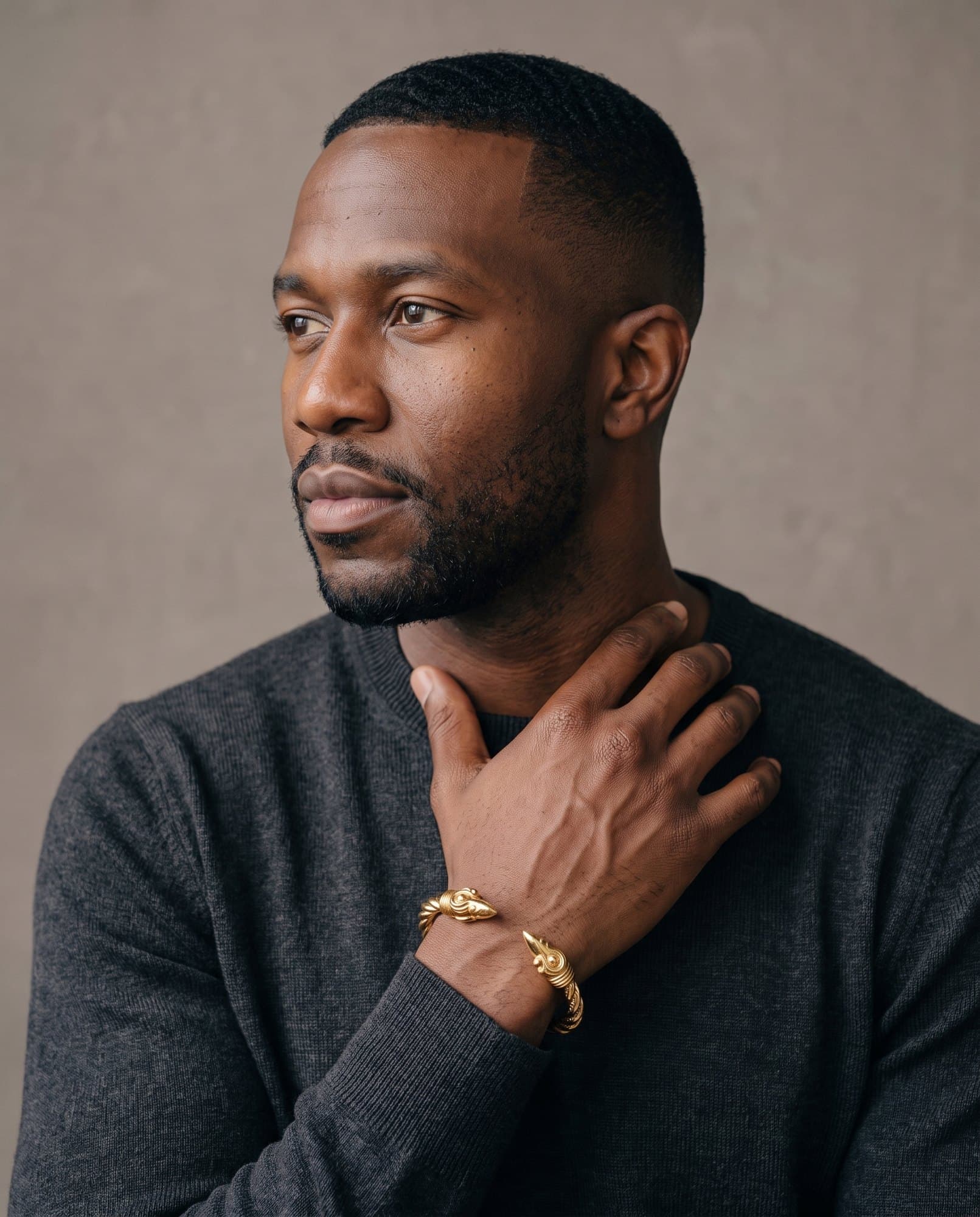 A man in a charcoal knit with his hand at his collarbone, the Horus Falcon Cuff on his wrist.