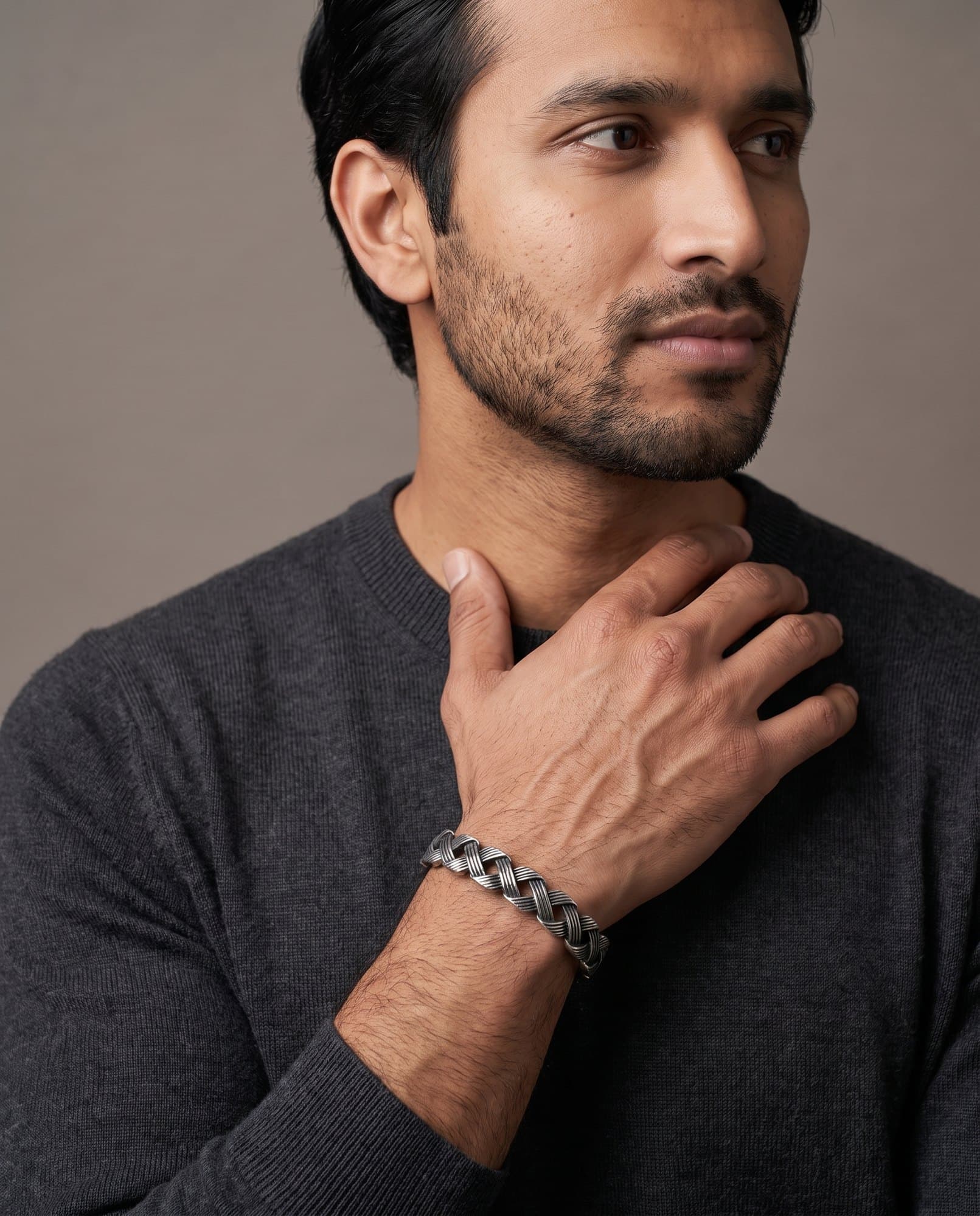 A man in a charcoal knit with his hand at his collarbone, the Horus Lattice Cuff on his wrist.