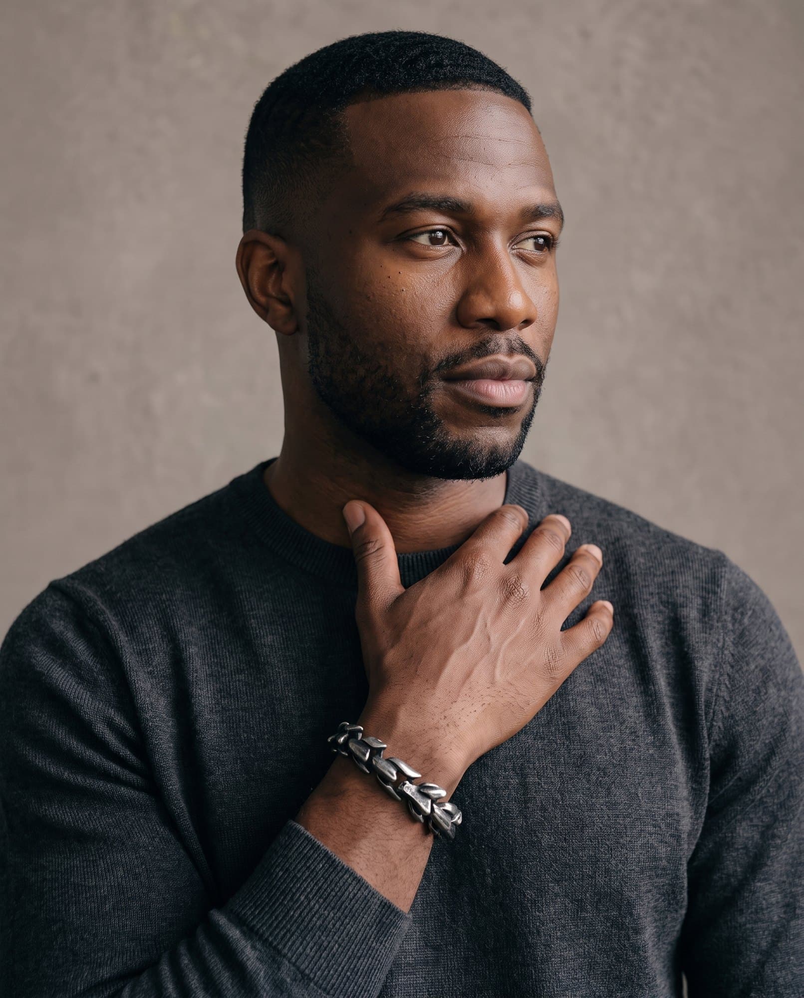 A man in a charcoal knit with his hand at his collarbone, the Horus Link Chain on his wrist.