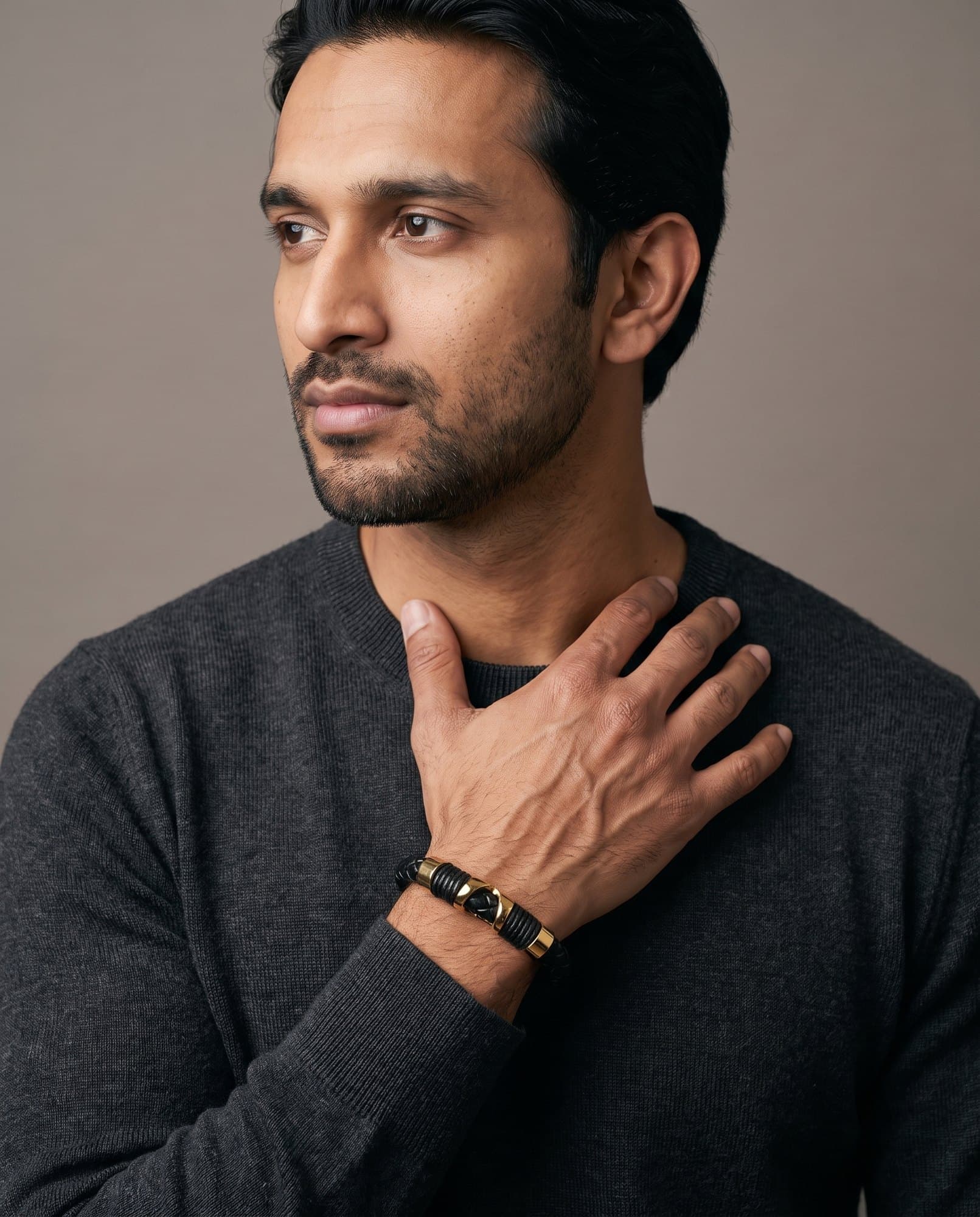 A man in a charcoal knit with his hand at his collarbone, the Horus Saddle Cord on his wrist.