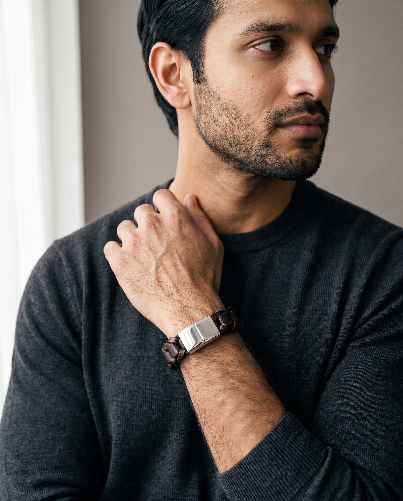 A man in a charcoal knit with his hand at his collarbone, the Interlaced Plait Cord on his wrist.