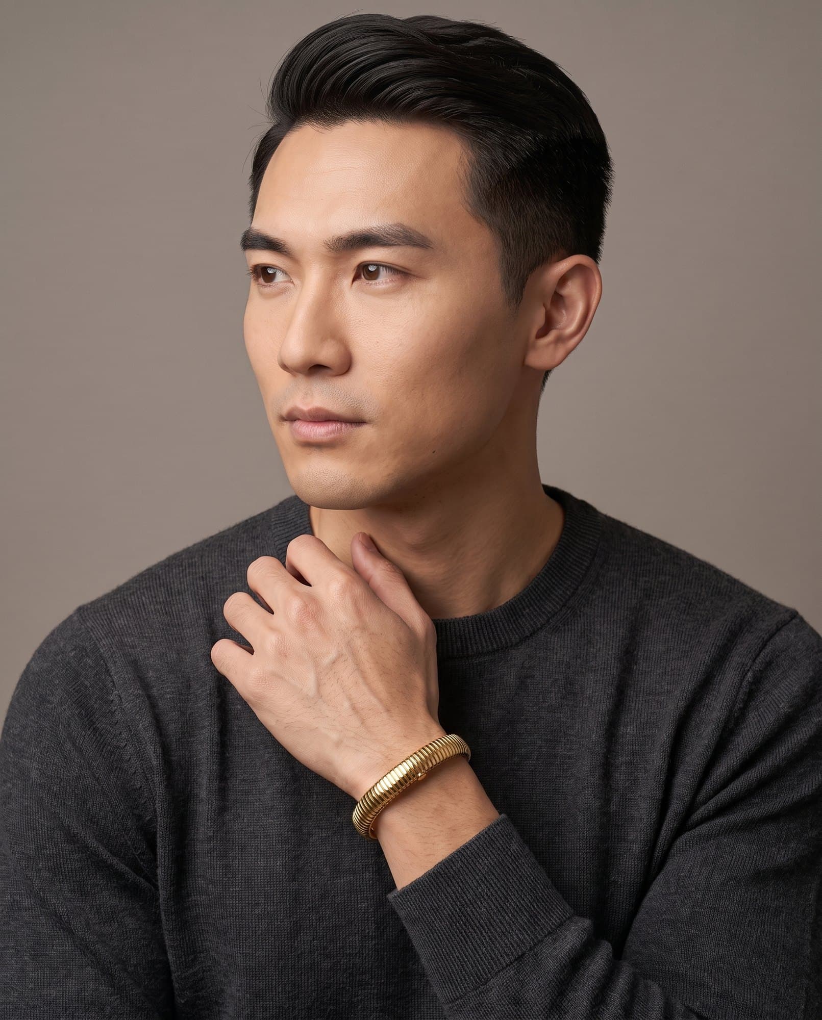 A man in a charcoal knit with his hand at his collarbone, the Luxor Coil Bangle on his wrist.
