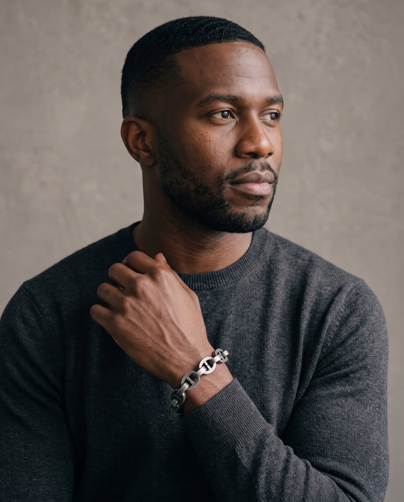 A man in a charcoal knit with his hand at his collarbone, the Mariner Chain on his wrist.