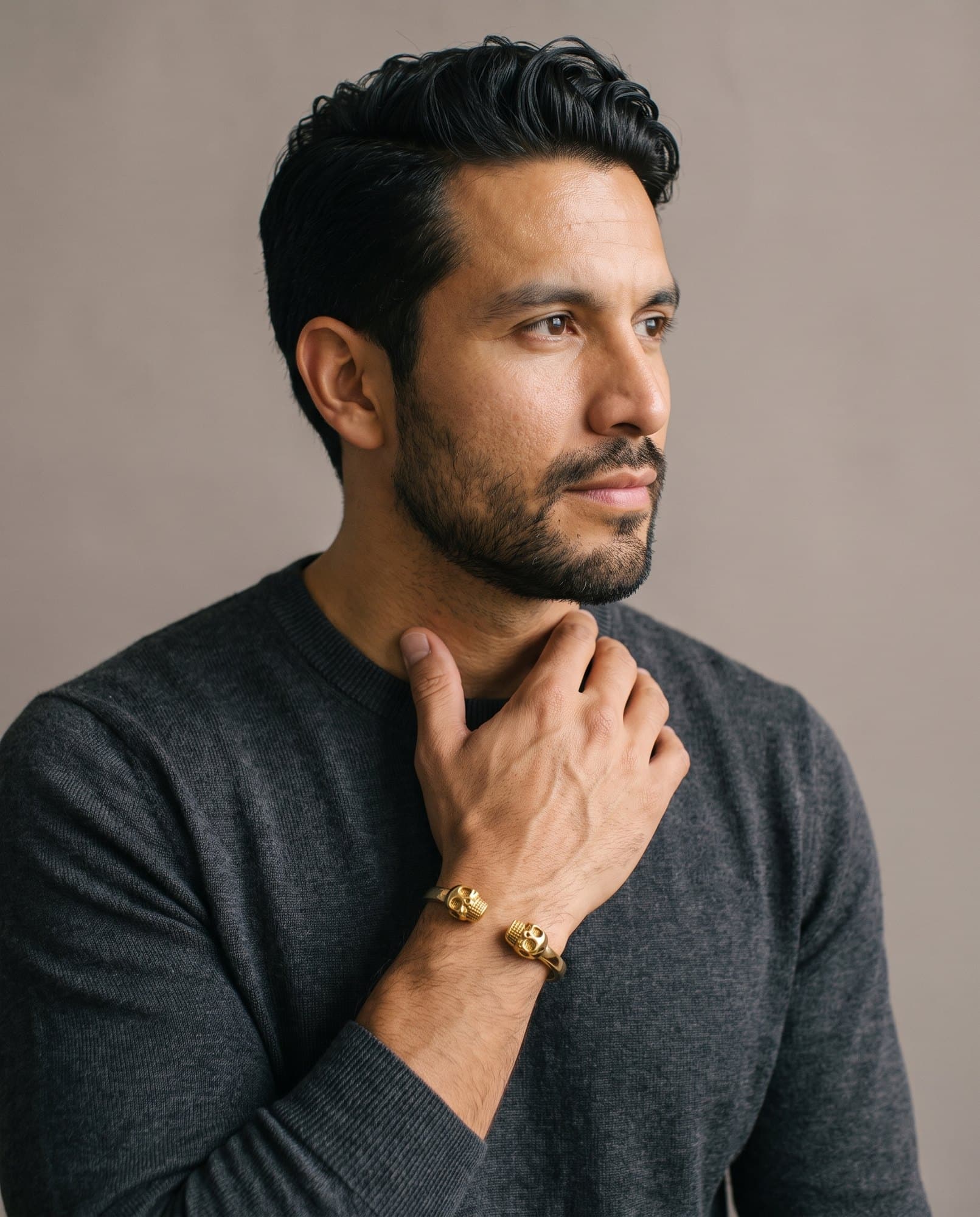 A man in a charcoal knit with his hand at his collarbone, the Pharaoh Skull Cuff on his wrist.