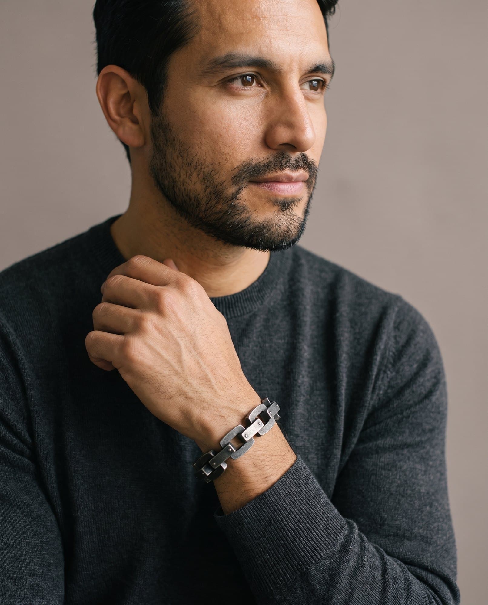 A man in a charcoal knit with his hand at his collarbone, the Rivet Block Chain on his wrist.