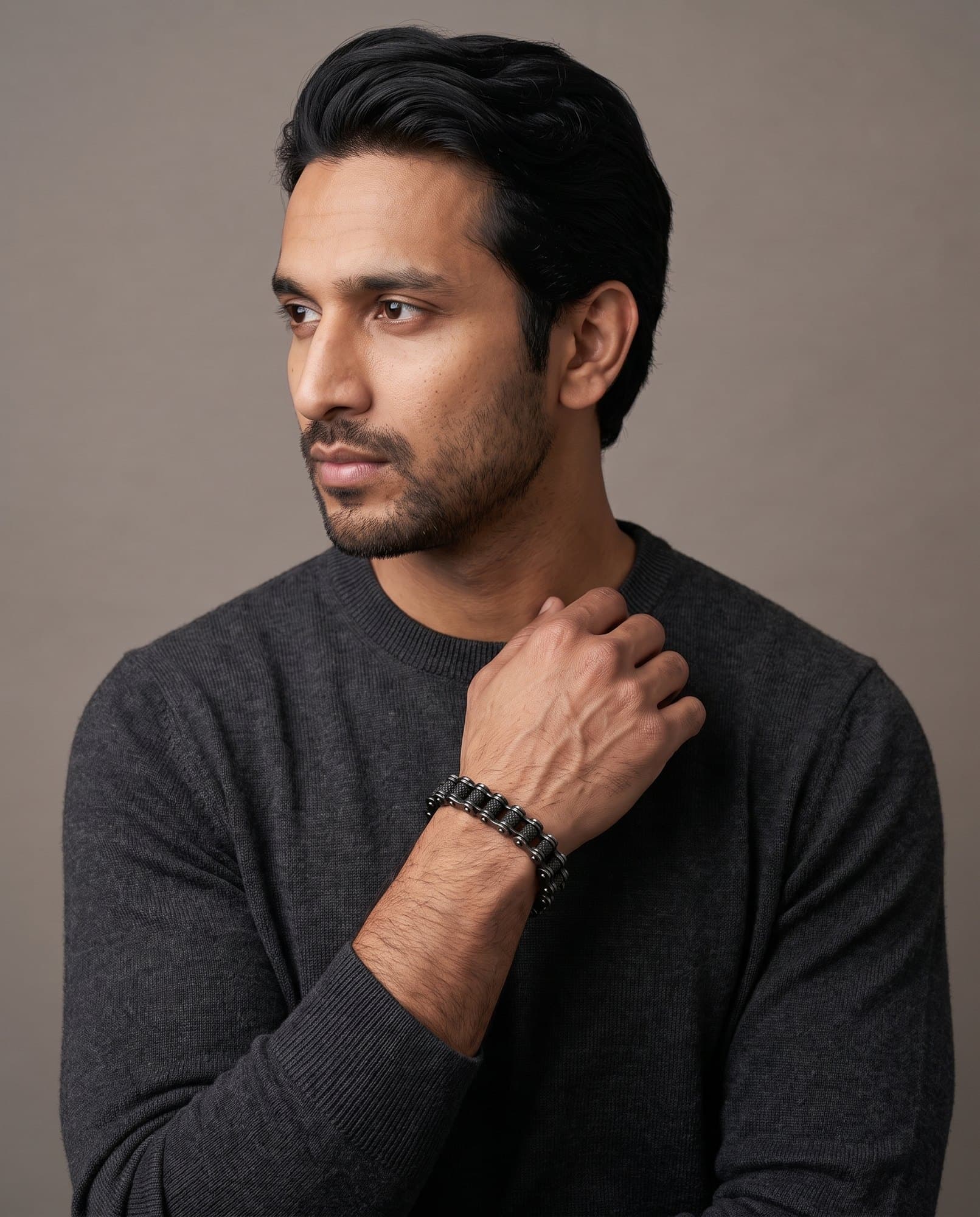 A man in a charcoal knit with his hand at his collarbone, the Roller Chain on his wrist.