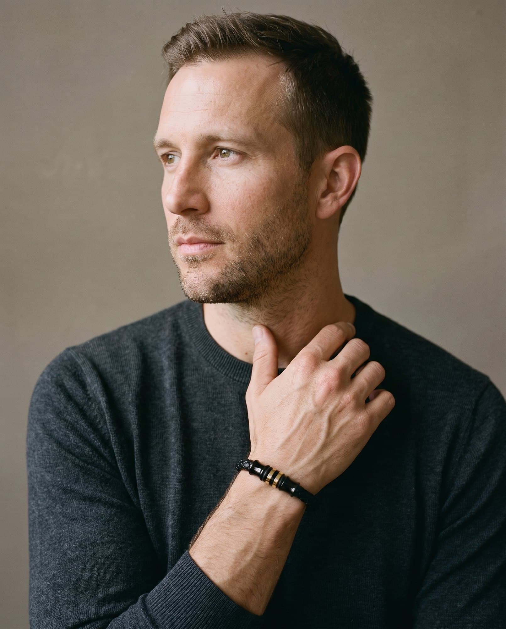 A man in a charcoal knit with his hand at his collarbone, the Scarab Banded Cord on his wrist.