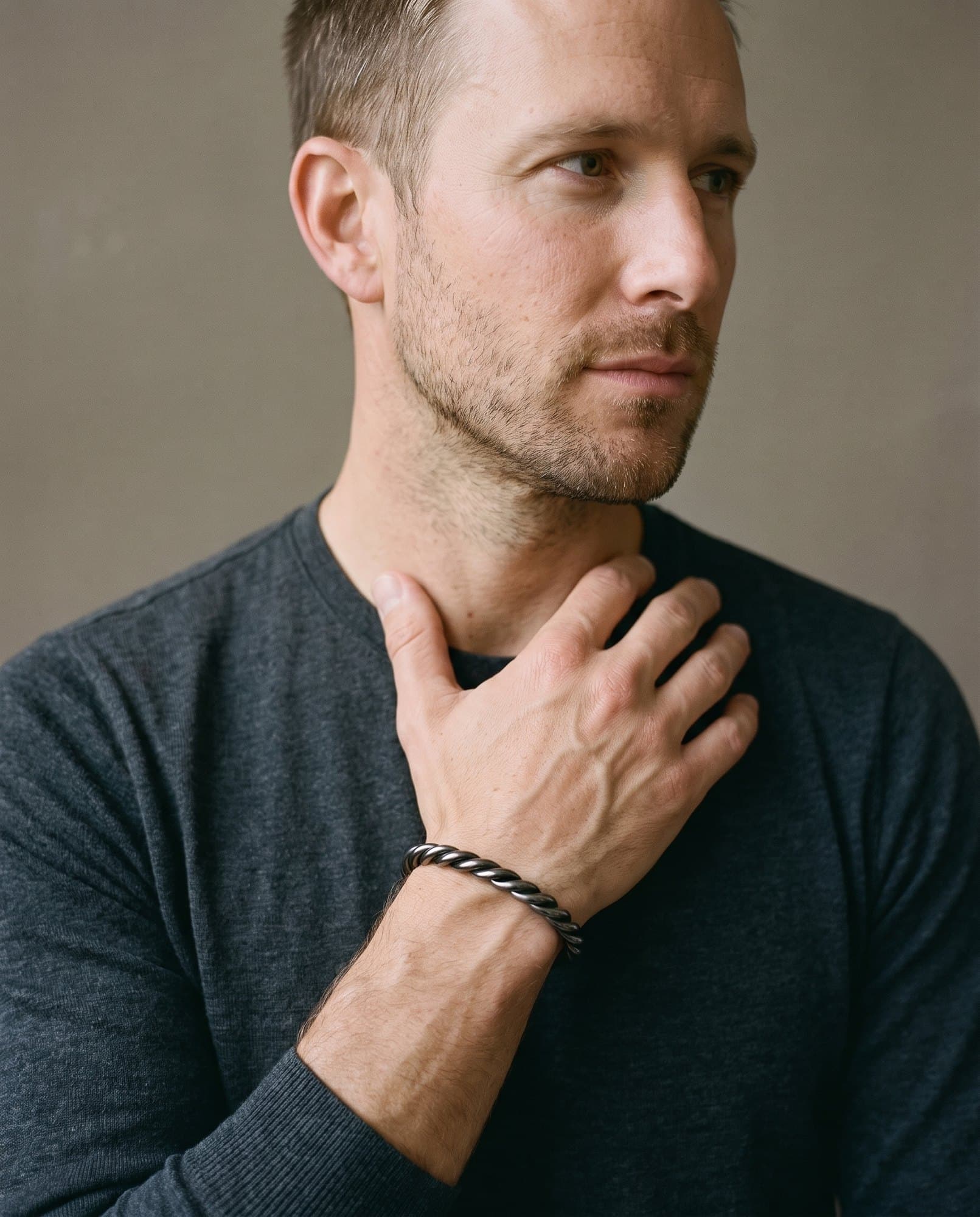 A man in a charcoal knit with his hand at his collarbone, the Scribe Rope Cuff on his wrist.