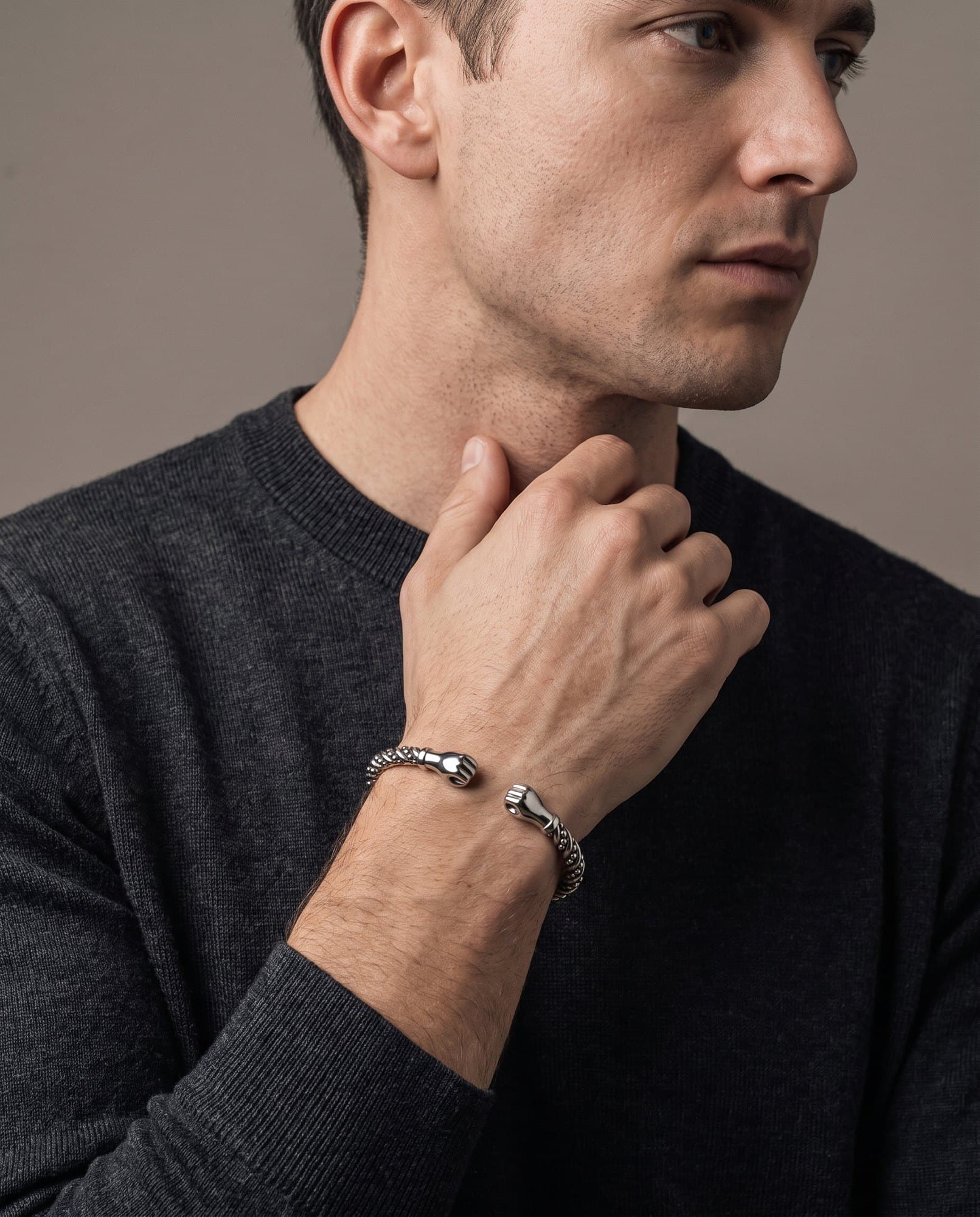 A man in a charcoal knit with his hand at his collarbone, the Sovereign Fist Cuff on his wrist.