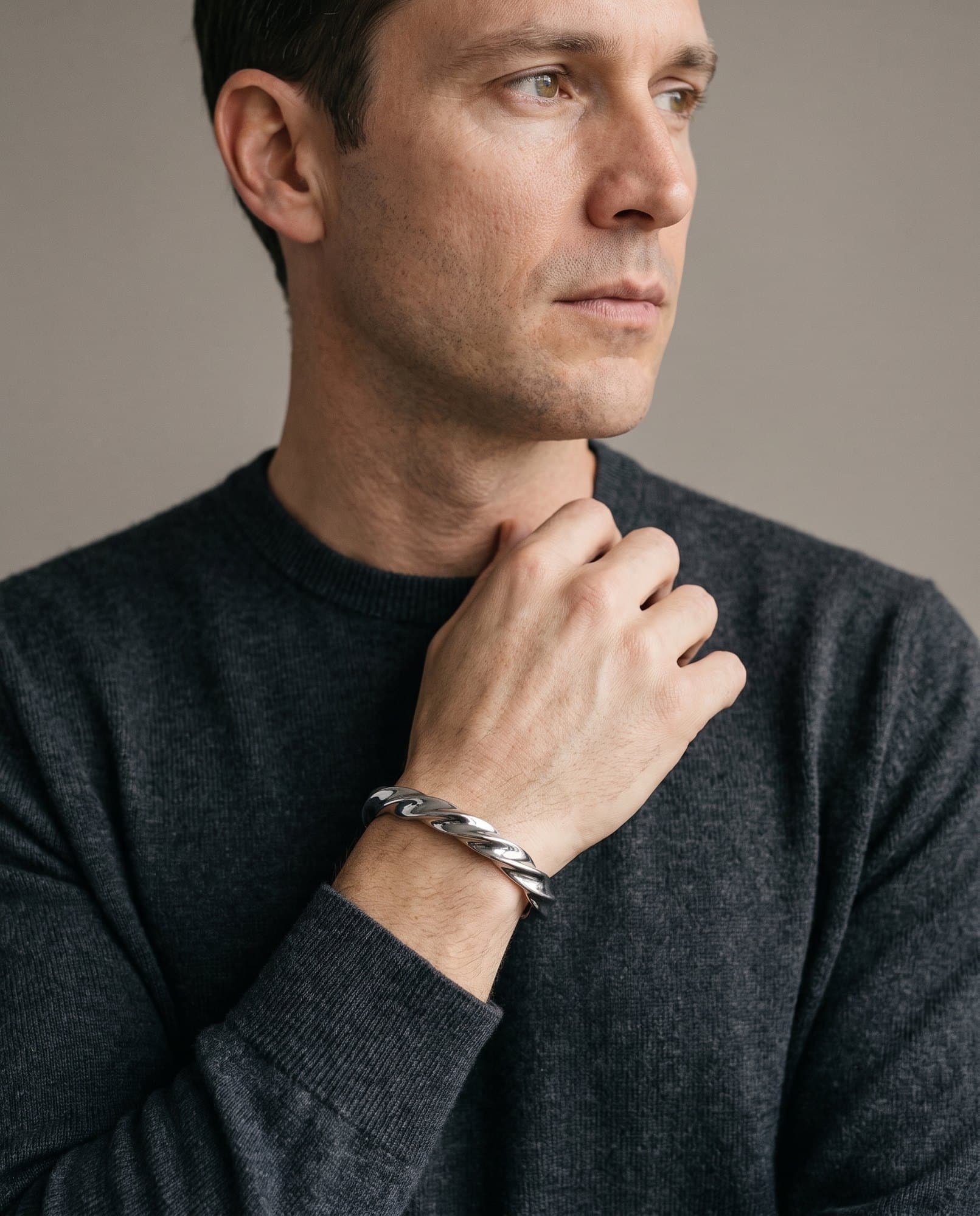 A man in a charcoal knit with his hand at his collarbone, the Sphinx Twist Cuff on his wrist.