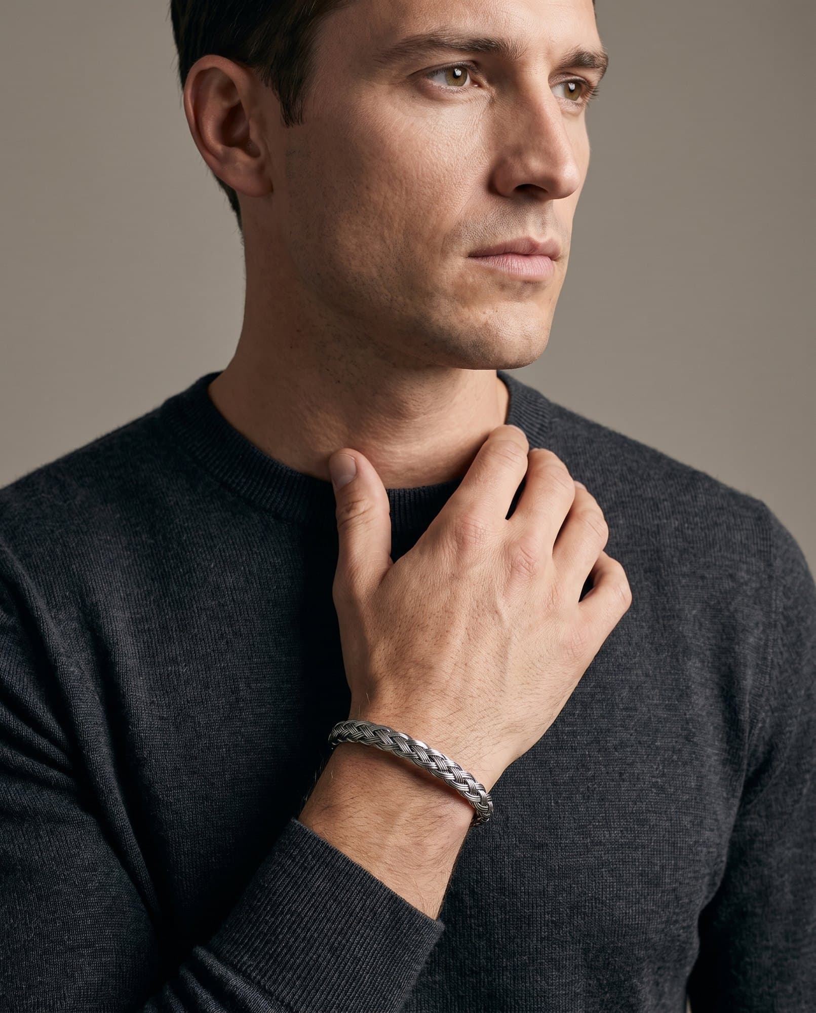 A man in a charcoal knit with his hand at his collarbone, a woven steel cuff on his wrist.