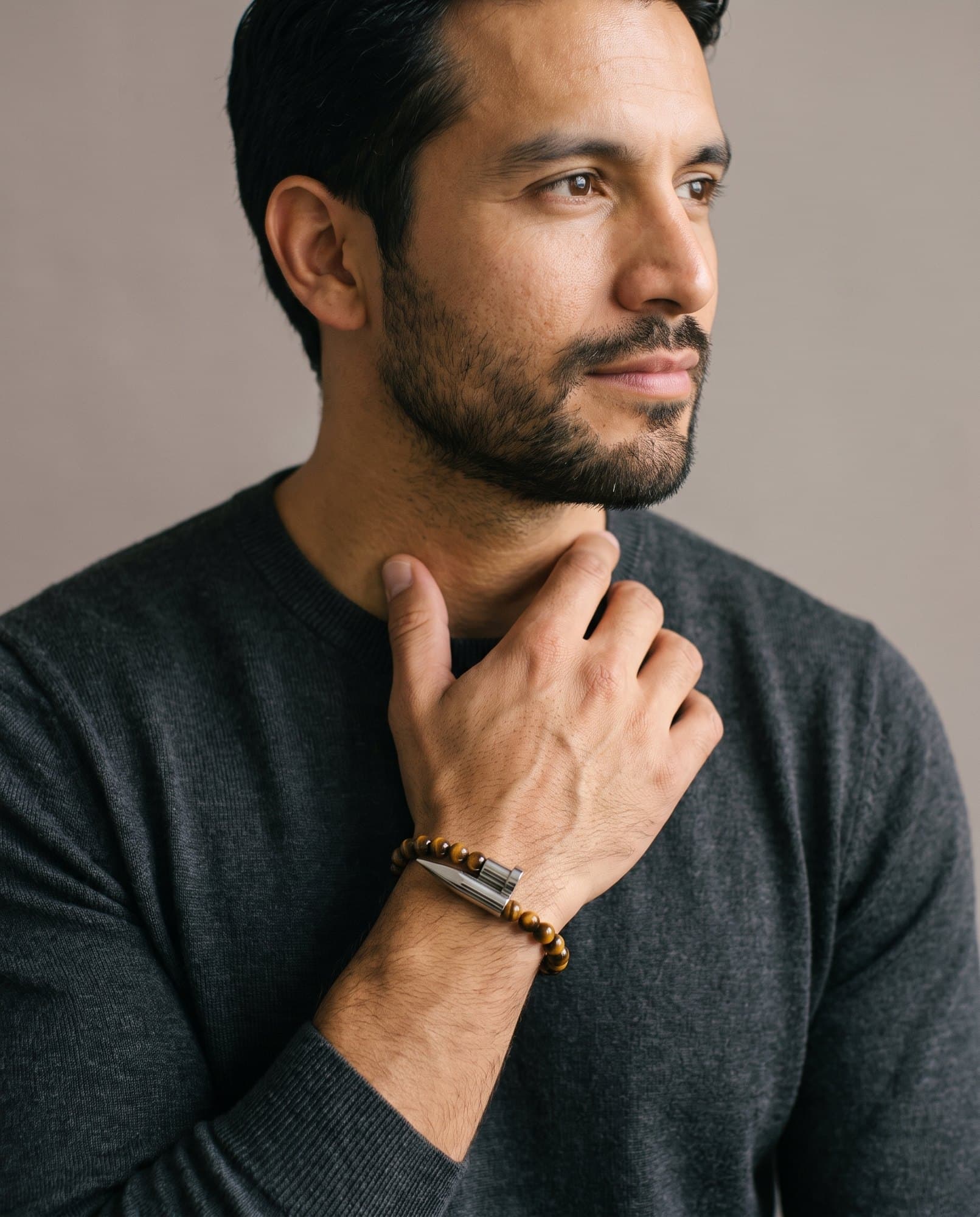 A man in a charcoal knit with his hand at his collarbone, the Tiger Spike Bracelet on his wrist.
