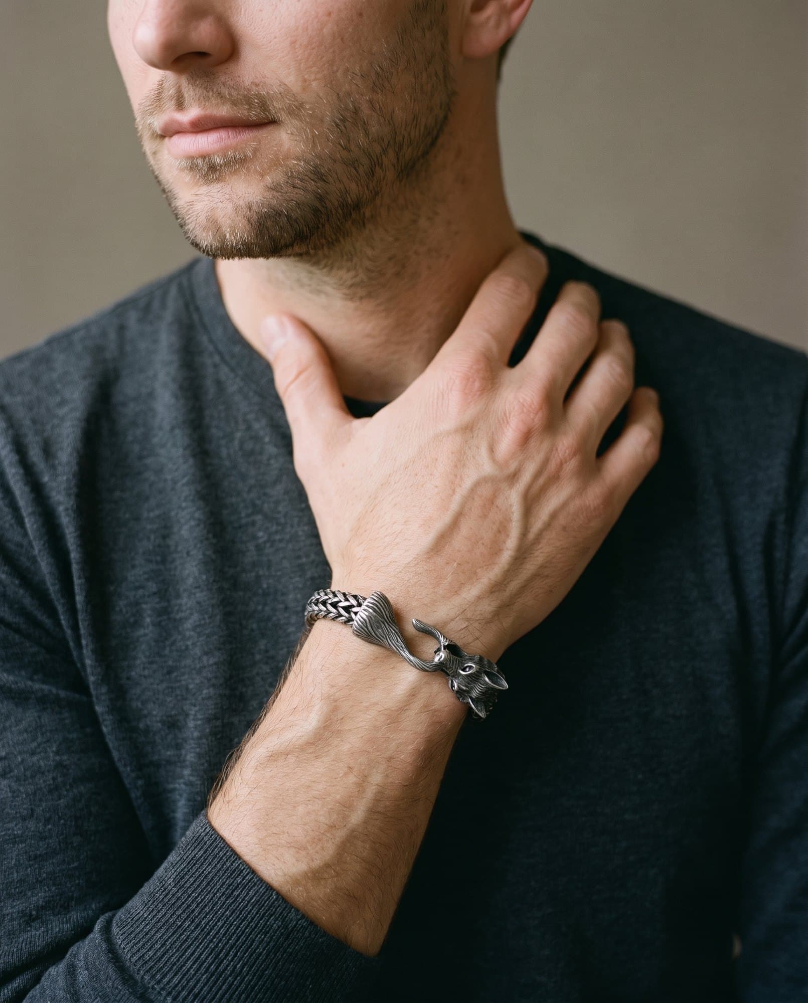 A man in a charcoal knit with his hand at his collarbone, the Wolf Head Chain on his wrist.