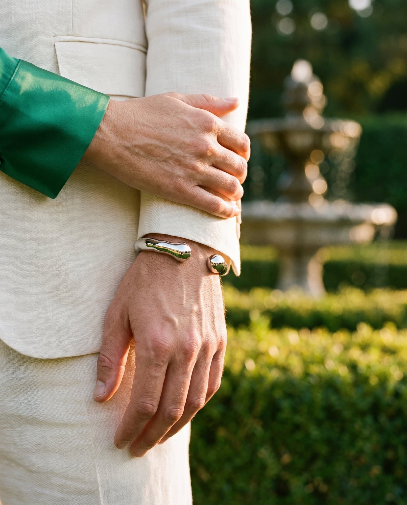A woman's hand resting lightly on a man's wrist above a mirror polished steel cuff, a fountain soft behind.