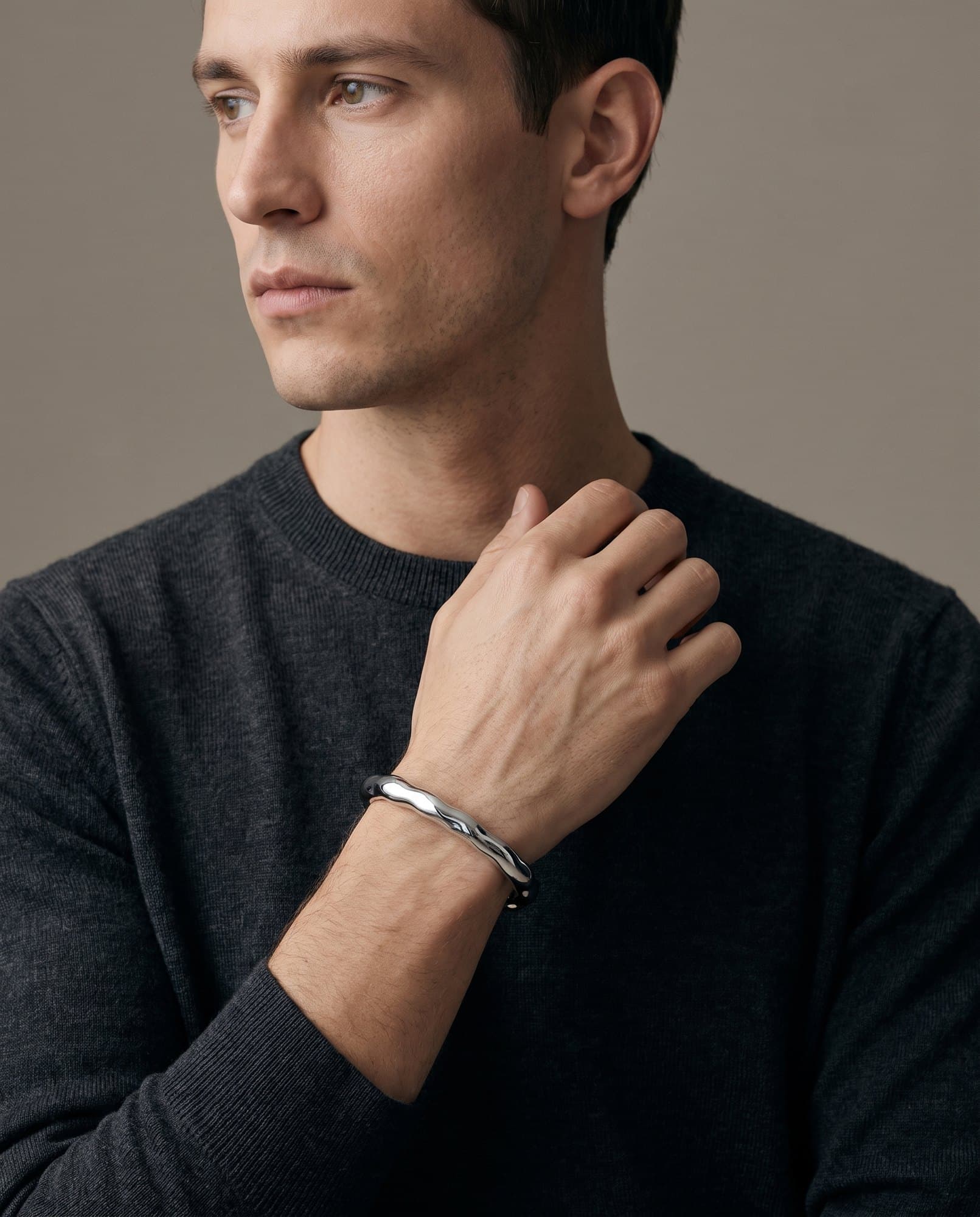 A man in a charcoal knit with his hand at his collarbone, the Nile Molten Cuff on his wrist.