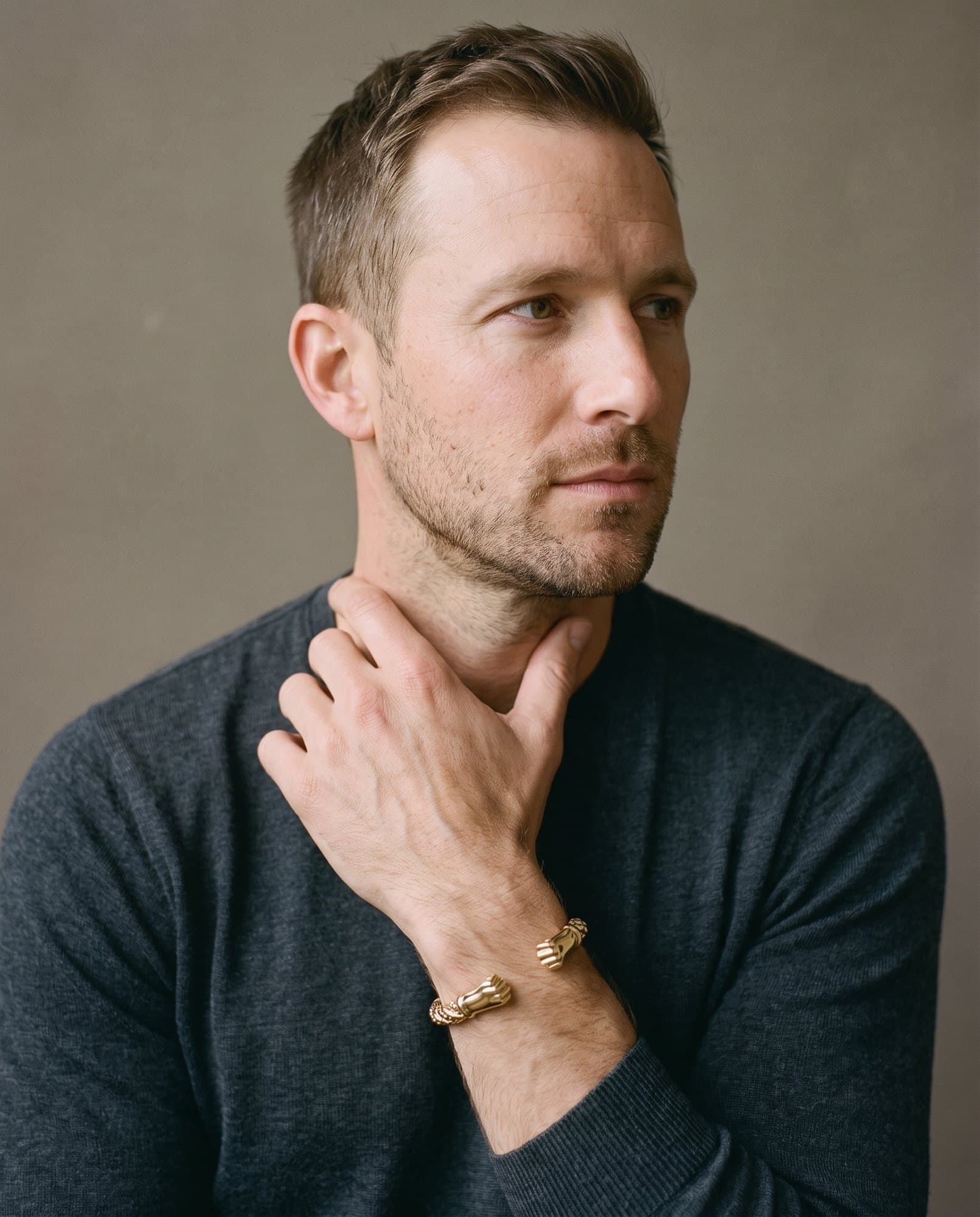 A man in a charcoal knit with his hand at his collarbone, the Pharaoh Fist Cuff on his wrist.