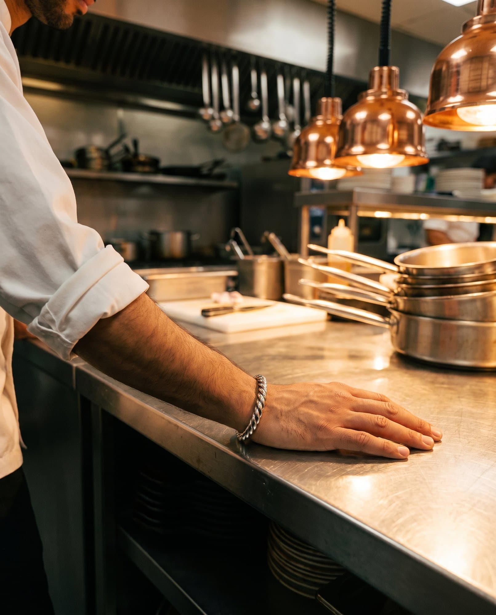 A strong hand resting flat on a steel kitchen counter, a broad twisted steel cuff at the wrist.