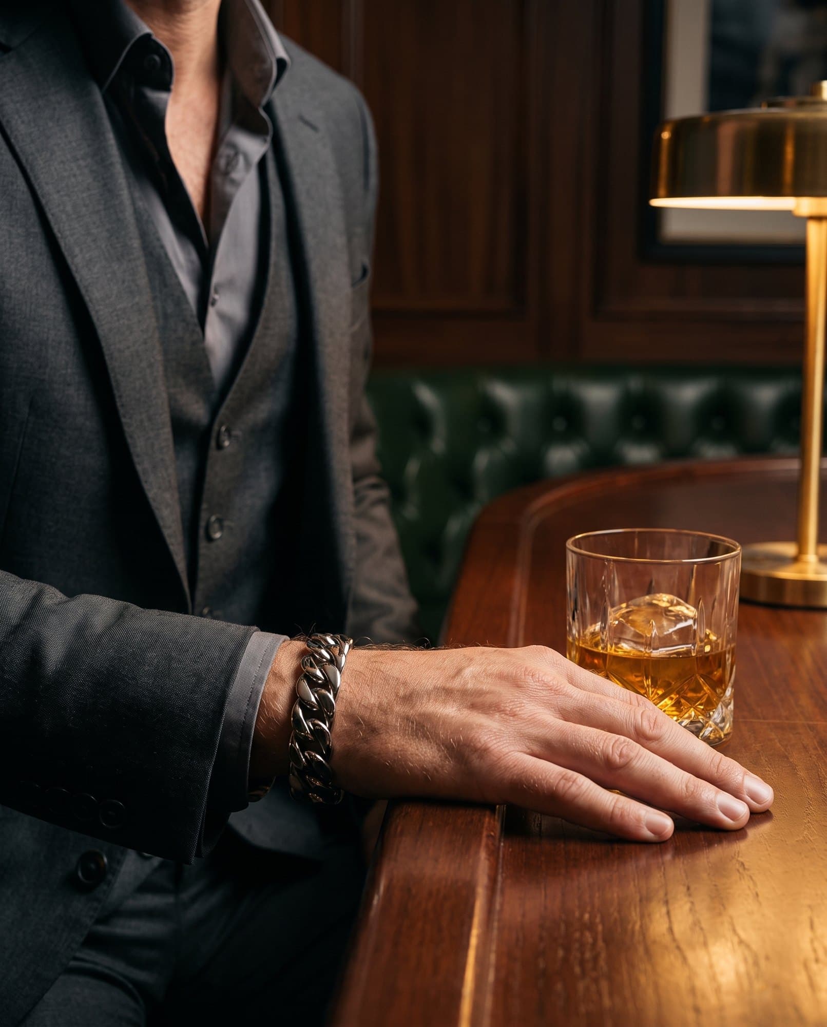 A forearm resting on a polished wood bar beside a whisky tumbler, a heavy curb link steel cuff at the wrist.