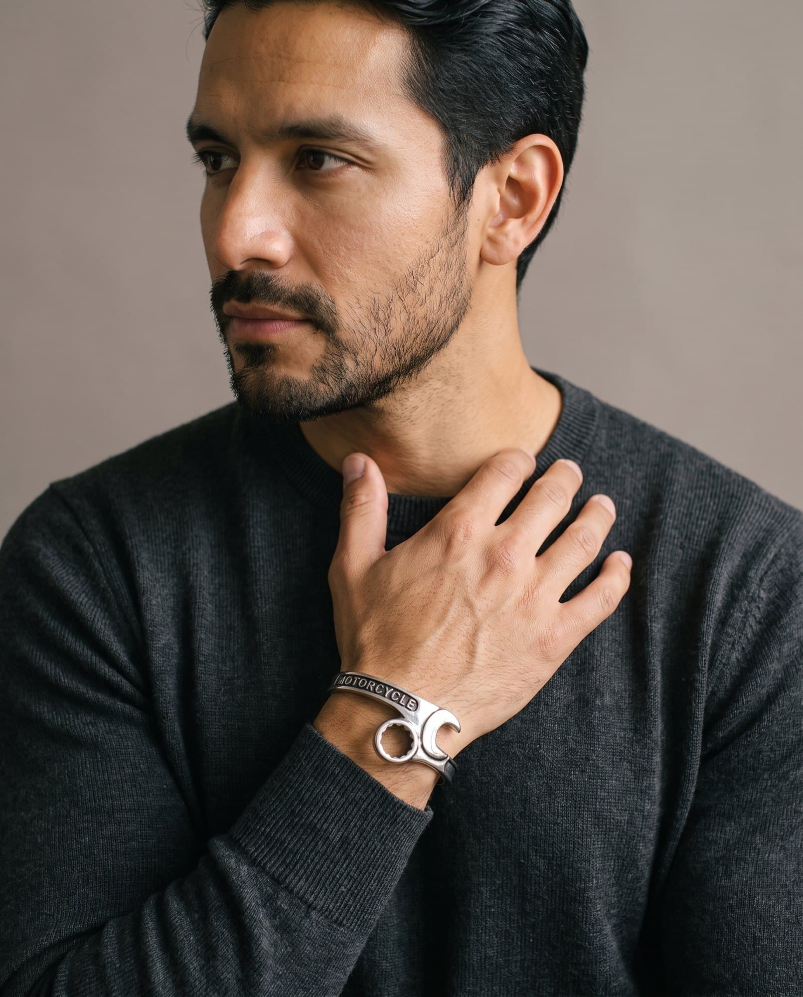 A man in a charcoal knit with his hand at his collarbone, the Piston Wheel Cuff on his wrist.