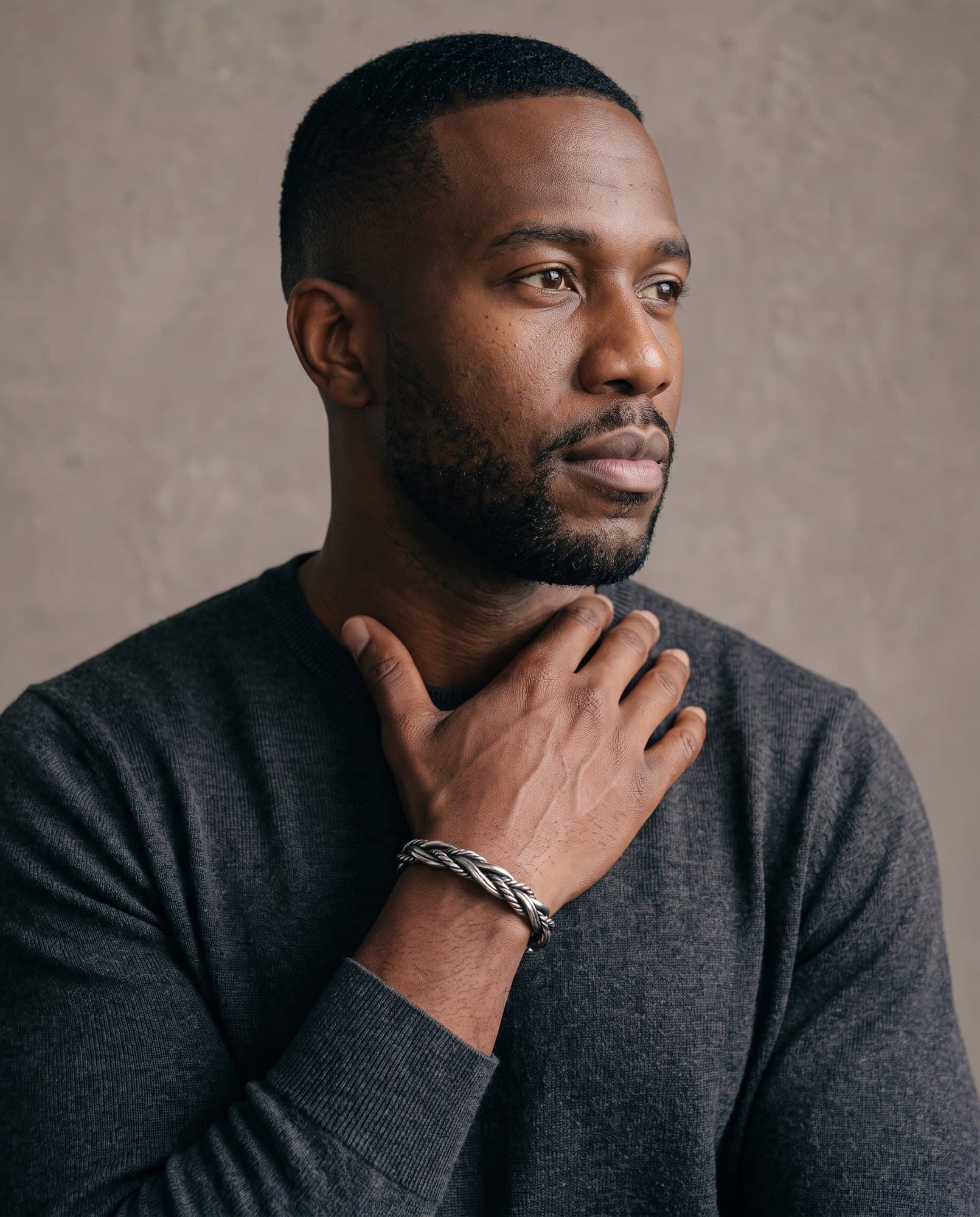 A man in a charcoal knit with his hand at his collarbone, the Sovereign Braid Cuff on his wrist.