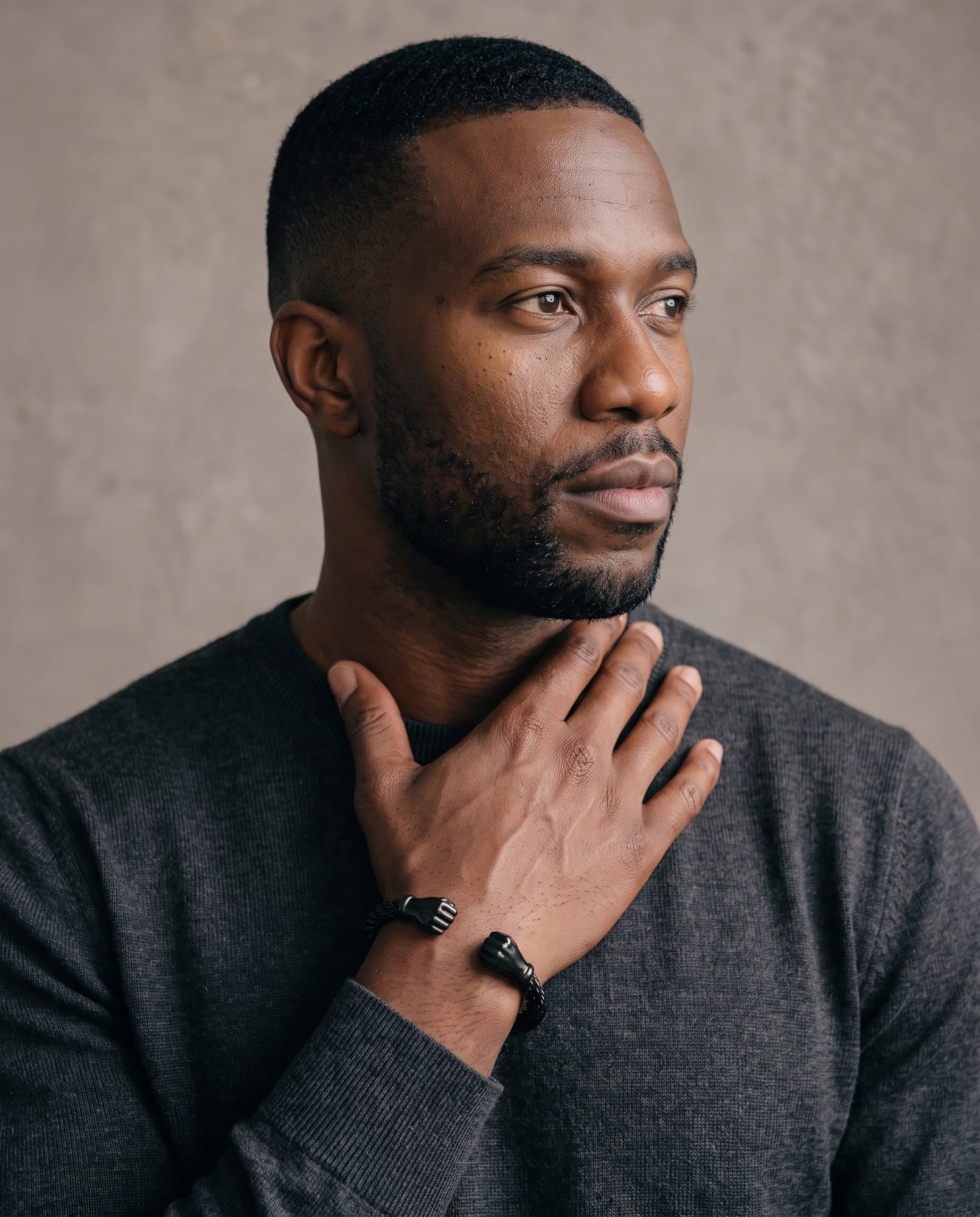 A man in a charcoal knit with his hand at his collarbone, the Sphinx Fist Cuff on his wrist.