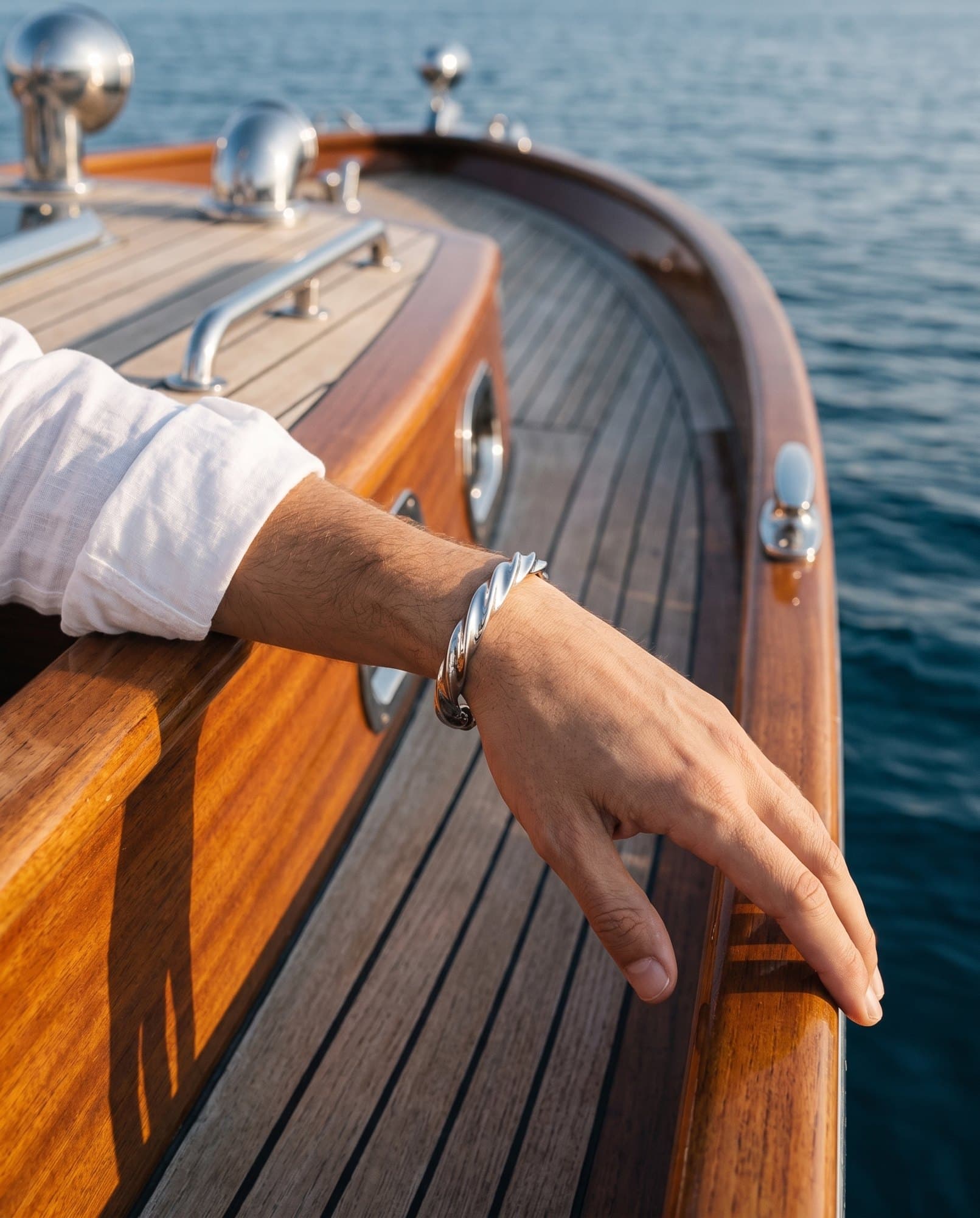 A forearm resting on a varnished teak rail over the sea, a heavy twisted steel cuff at the wrist.