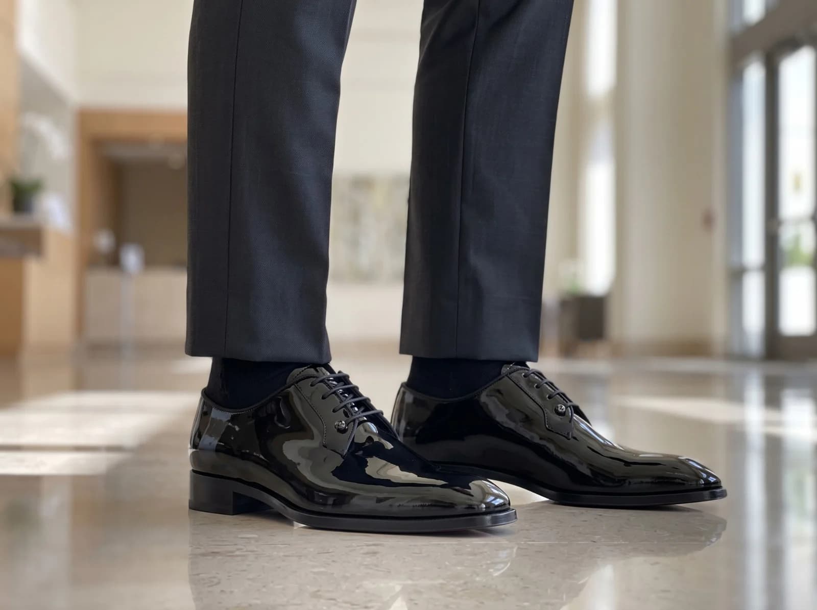 Patent derbys worn in a hotel lobby with grey trousers and black socks